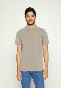 Camisa polo bege de mangas curtas, com colarinho e placket de dois botões. Usada com jeans azuis, contra um fundo simples.