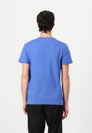 Mężczyzna o ciemnych włosach ubrany w prosty niebieski t-shirt z krótkim rękawem i czarne spodnie, stojący tyłem na białym tle.