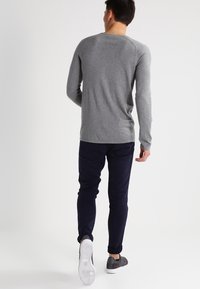 Chemise grise à manches longues en coton, coupe ajustée, associée à un pantalon bleu foncé et des baskets noires, vue de dos.