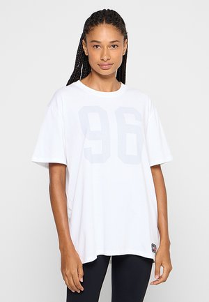 Femme aux cheveux tressés portant un t-shirt blanc ample avec un "96" gris clair à l'avant et des leggings noirs, debout devant un fond uni.