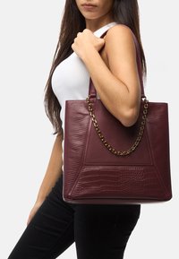 Borsa tote in ecopelle color bordeaux con dettagli testurizzati, un design geometrico e manico in catena dorata. Forma liscia e strutturata.