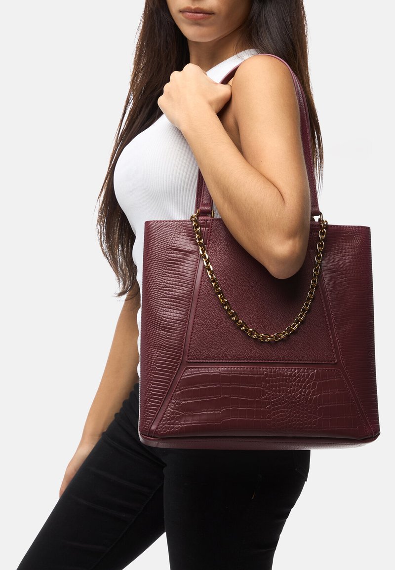 Borsa tote in ecopelle color bordeaux con dettagli testurizzati, un design geometrico e manico in catena dorata. Forma liscia e strutturata.