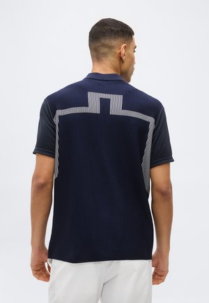 Hombre con camisa azul marino de manga corta con patrón de rayas geométricas blancas en la parte posterior y pantalones blancos, de espaldas contra un fondo liso.