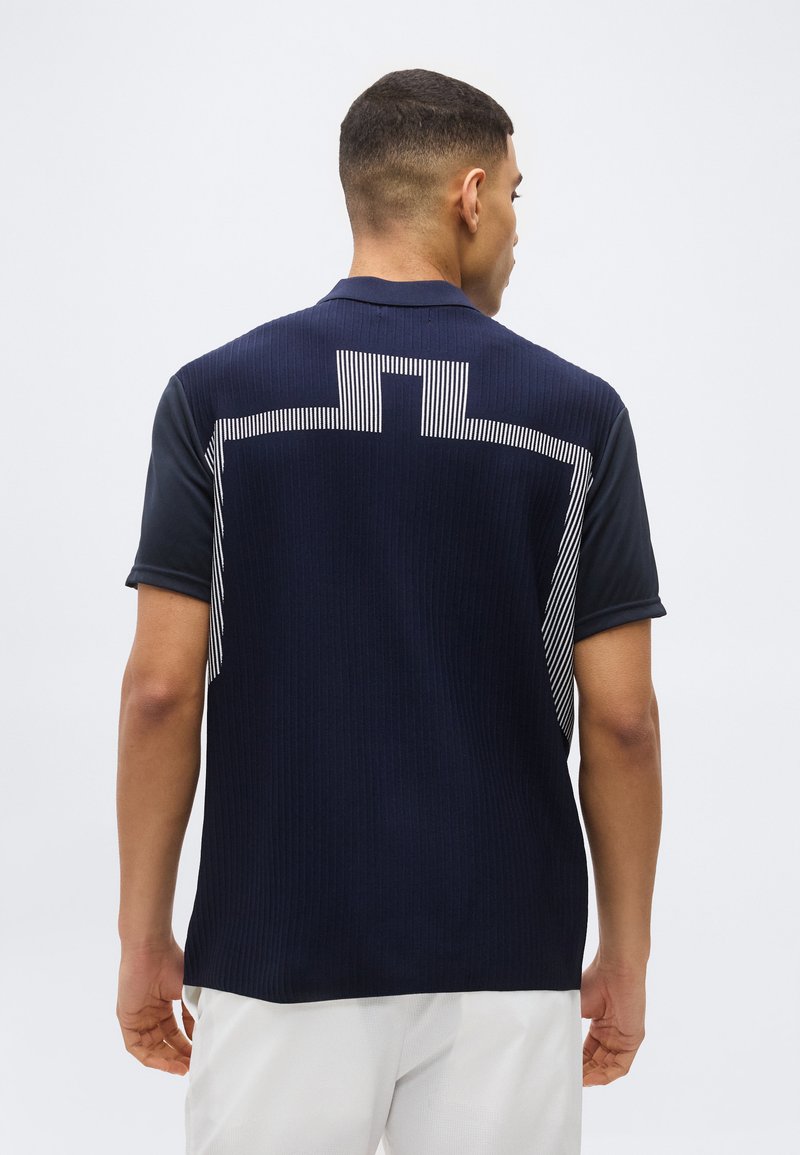 Άνδρας που φοράει μπλε ναυτικό κοντομάνικο πουκάμισο με λευκό γεωμετρικό μοτίβο ρίγες στην πλάτη και λευκό παντελόνι, γυρισμένος πλάτη σε απλό φόντο.