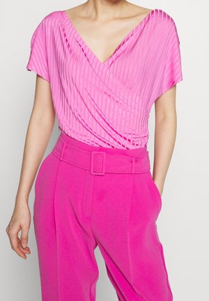 Femme portant un top cache-cœur plissé rose vif associé à un pantalon rose taille haute ceinturé, debout devant un fond uni.
