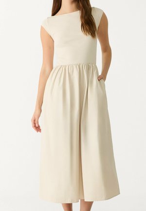 Mujer que lleva un vestido midi beige claro sin mangas con la parte superior ajustada y falda fruncida, una mano apoyada en un bolsillo lateral.