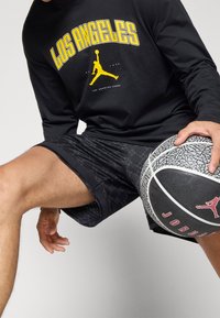 Person wearing black Los Angeles long-sleeve shirt and black shorts, holding a black and white Jordan basketball in mid-dribble stance.