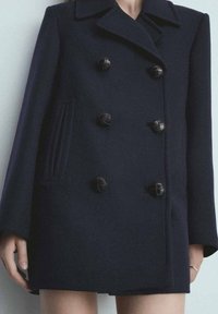 Merineuleinen kaksirivinen takki, jossa on leveä kaulus, isot mustat napit ja sivutaskut, joissa on pystysuuntaista veteen liittyvää yksityiskohtaa.