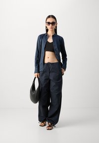 Camicia a button-up blu navy, top corto nero e pantaloni larghi blu navy con coulisse. Borsa nera e sandali piatti con cinturini incrociati.