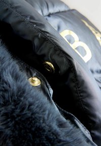 Svart vadderad jacka med guldfärgad knapprade och en mjuk, fluffig krage. Tyget är slätt med quiltad struktur och djärva bokstäver.