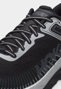 Gros plan sur une chaussure de course noire et grise avec une tige en mesh, des lacets à motifs et la marque "HOKA" sur le côté et la languette.