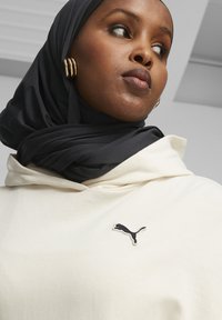Creme hoodie med snor, prydet med et sort broderet Puma-logo. Matchet med et mørkt, tekstureret hijab og guld hoop-øreringe.