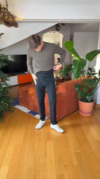 Bărbat cu pulover maro ajustând o pereche de pantaloni închiși la culoare în camera de zi cu o canapea portocalie, o plantă mare verde, un brad de Crăciun și podea din lemn.