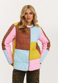 Žena s blond vlasmi, oblečená v prešívanej, viacfarebnej patchworkovej bunde a nohaviciach s leopardím vzorom, stojí so zatajenými rukami v vreckách.