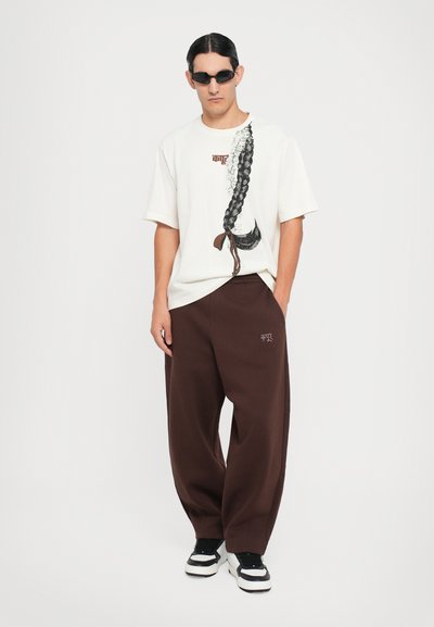 Hellfarbener, übergroßer T-Shirt mit einem geflochtenen Grafikdesign; braune Jogginghose; schwarz-weiße Sneaker; minimalistischem Stil.