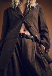 Hnedé sako s prúžkami a dvoma gombíkmi, kombinované s vysokými nohavicami a čiernym opaskom. Textúra látky je hladká.