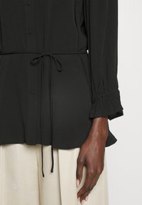 Blouse noire à manches longues avec boutons, taille à cordon et poignets élastiques à volants portée sur un pantalon beige clair sur un mannequin à peau foncée.