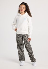 Ragazza giovane con capelli ricci che indossa un hoodie bianco e pantaloni con stampa leopardata, in piedi con una mano in tasca e scarpe da ginnastica bianche.