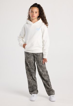 Jeune fille aux cheveux bouclés, portant un sweat à capuche blanc et un pantalon en imprimé léopard, se tenant avec une main dans la poche et des baskets blanches.