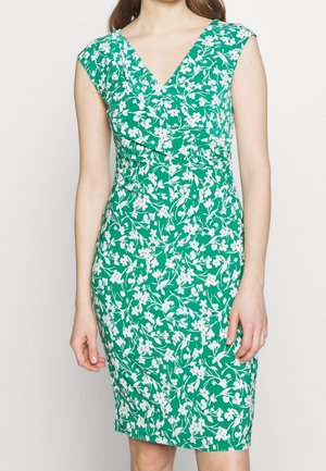 Ärmelloses grünes Kleid mit weißem Blumenmuster, V-Ausschnitt und taillierter Taille, getragen von einer stehenden Person vor einem einfarbigen Hintergrund.
