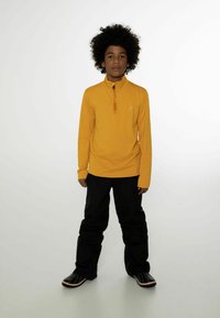 Gelbes Langarm-Half-Zip-Oberteil mit Logo, kombiniert mit schwarzen Hosen und schwarzen Schuhen mit roten Akzenten. Glatte Stofftextur.
