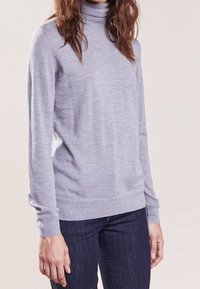 Hellgrauer Rollkragenpullover aus einem weichen, strukturierten Stoff mit langen Ärmeln und einem enganliegenden Saum, kombiniert mit dunklen Jeans, vor einem neutralen Hintergrund gezeigt.