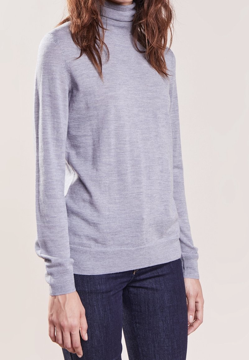 Hellgrauer Rollkragenpullover aus einem weichen, strukturierten Stoff mit langen Ärmeln und einem enganliegenden Saum, kombiniert mit dunklen Jeans, vor einem neutralen Hintergrund gezeigt.
