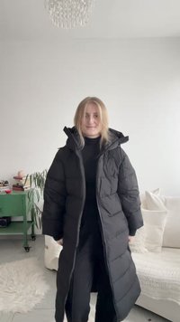 Casaco acolchoado preto com design longo e oversize, com capuz e fecho de correr. O tecido tem uma textura lisa e brilhante.