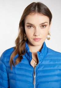 Giacca puffer blu con colletto alto, cuciture orizzontali e zip frontale. Zip metallica lucida e tessuto testurizzato ben visibili.