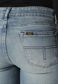 Lyseblå denimbukser med en tettsittende silhuett, fremtredende sømmer og en liten svart etikett med teksten "Lois" på baklommen.