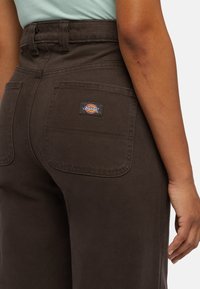 Hnědé kalhoty s vysokým pasem vyrobené z texturované látky, s dvojitými zadními kapsami a černou nášivkou s logem Dickies na pravé kapse.