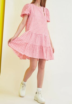 Femme portant une robe à manches bouffantes à carreaux roses et blancs et des baskets blanches, tenant l'ourlet de la robe tout en se tenant devant un fond uni.