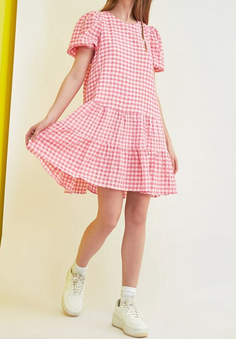 Femme portant une robe à manches bouffantes à carreaux roses et blancs et des baskets blanches, tenant l'ourlet de la robe tout en se tenant devant un fond uni.