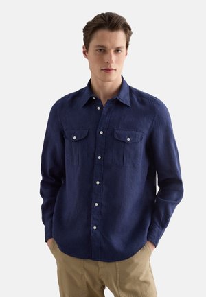 Giovane uomo con capelli castani corti che indossa una camicia blu navy con bottoni e tasche sul petto e pantaloni beige su sfondo semplice.