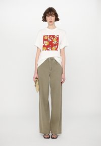 Weißes T-Shirt mit einem roten floralen Grafikdesign, kombiniert mit olivgrünen weit geschnittenen Hosen. Das Model hält eine goldene Clutch und trägt schwarze Sandalen.