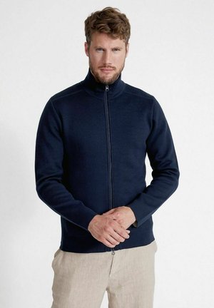 Marineblå lynlåsjakke i blødt stof, med høj krave og glat tekstur. Designet er tætsiddende med lange ærmer og en minimalistisk stil.