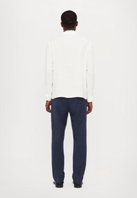 Cămașă albă cu mâneci lungi și hem curbat, asociată cu pantaloni bleumarin. Materialul pare neted; cămașa are manșete cu nasturi și guler.