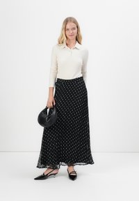 Lunga gonna nera plissettata con pois bianchi, abbinata a una maglietta a polo a maniche lunghe a coste chiare e scarpe nere con punta affusolata.