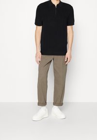 Černé polo tričko s krátkým rukávem a zipovým límcem, texturovaný pletený vzor. V kombinaci s ohrnutými béžovými kalhotami a bílými teniskami.