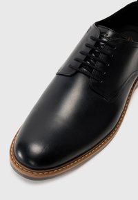 Zapato de vestir de cuero negro con un acabado suave, cierre de cordones en la parte frontal y suela de goma color tostado. Presenta una punta redondeada y discretos acentos de costura.