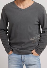 Graues Langarm-V-Ausschnitt-Shirt aus strukturiertem Stoff mit einem dezenten Logo auf der unteren rechten Seite, kombiniert mit hellblauen Jeans.