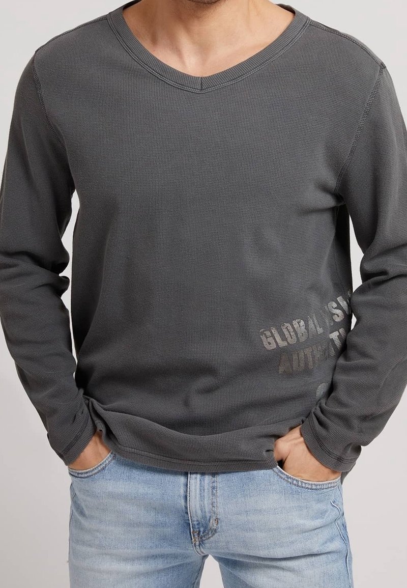 Camisa gris de manga larga y escote en V hecha de un tejido texturizado, con un sutil logo impreso en la parte inferior derecha, combinada con jeans de color azul claro.