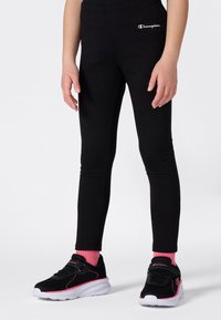 Niño vestido con leggings negros con el logo de Champion, calcetines rosas y zapatillas deportivas negras y rosas, de pie sobre un fondo blanco.