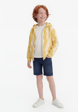 Παιδί με κόκκινα μαλλιά που στέκεται, φοράει κίτρινη και λευκή ριγέ μπλούζα με κουκούλα, λευκό t-shirt, σκούρο τζιν σορτς και λευκά αθλητικά παπούτσια, χαμογελώντας και κοιτάζοντας πλάγια.