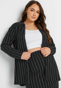 Camicia a righe bianco e nero con maniche lunghe, realizzata in tessuto testurizzato; indossata sopra un top corto bianco.