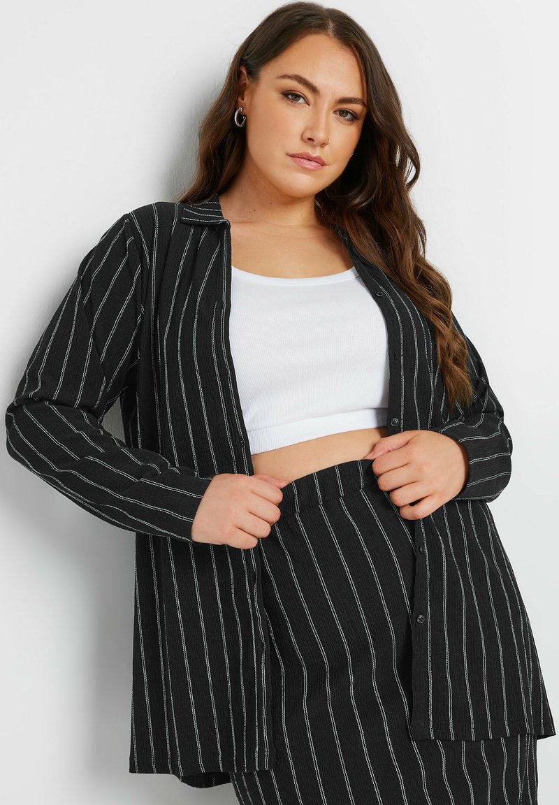 Camicia a righe bianco e nero con maniche lunghe, realizzata in tessuto testurizzato; indossata sopra un top corto bianco.