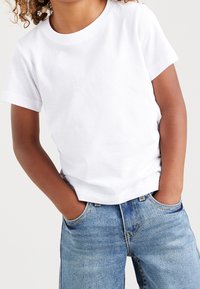Bambino che indossa una maglietta bianca a maniche corte e jeans blu, con le mani nelle tasche su uno sfondo bianco.