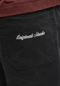 Poche arrière en denim noir avec texte brodé en blanc « Originals Studio » sous un ourlet de chemise marron.
