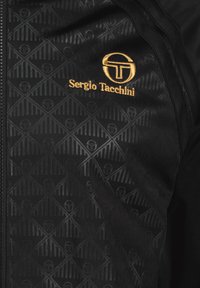 Sort zip-jacket med et struktureret geometrisk mønster, gul logo-broderi og en glat overflade.