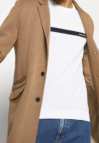 Brązowy płaszcz z mieszanki wełny z dwoma przednimi kieszeniami, jednorzędowym zapięciem i czarnymi guzikami. Noszony na białym swetrze w paski i dżinsach.